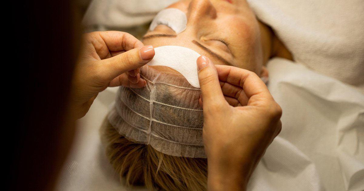 Beauty therapist performing facial treatment on client laying on bed covered with soft couch roll in a professional salon environment.
