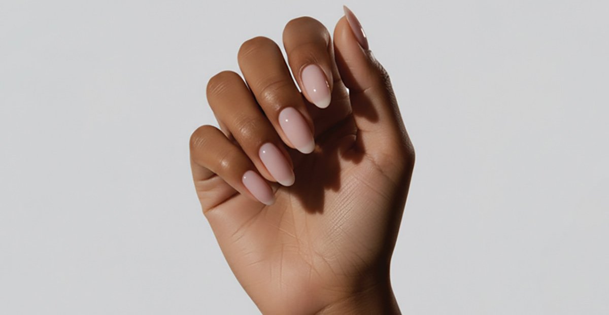 A hand featuring oval-shaped nails with a sheer, glossy milky-pink manicure against a light grey background.