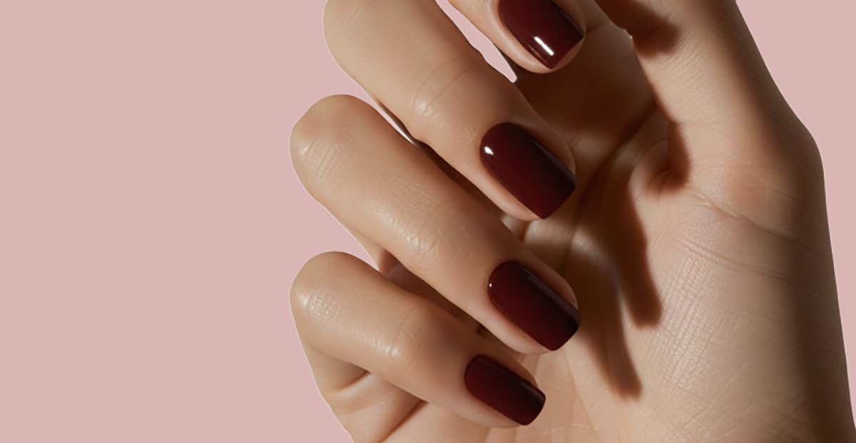 ATL TEXT A hand with short, square-shaped nails painted in a glossy, deep burgundy polish against a soft pink background.