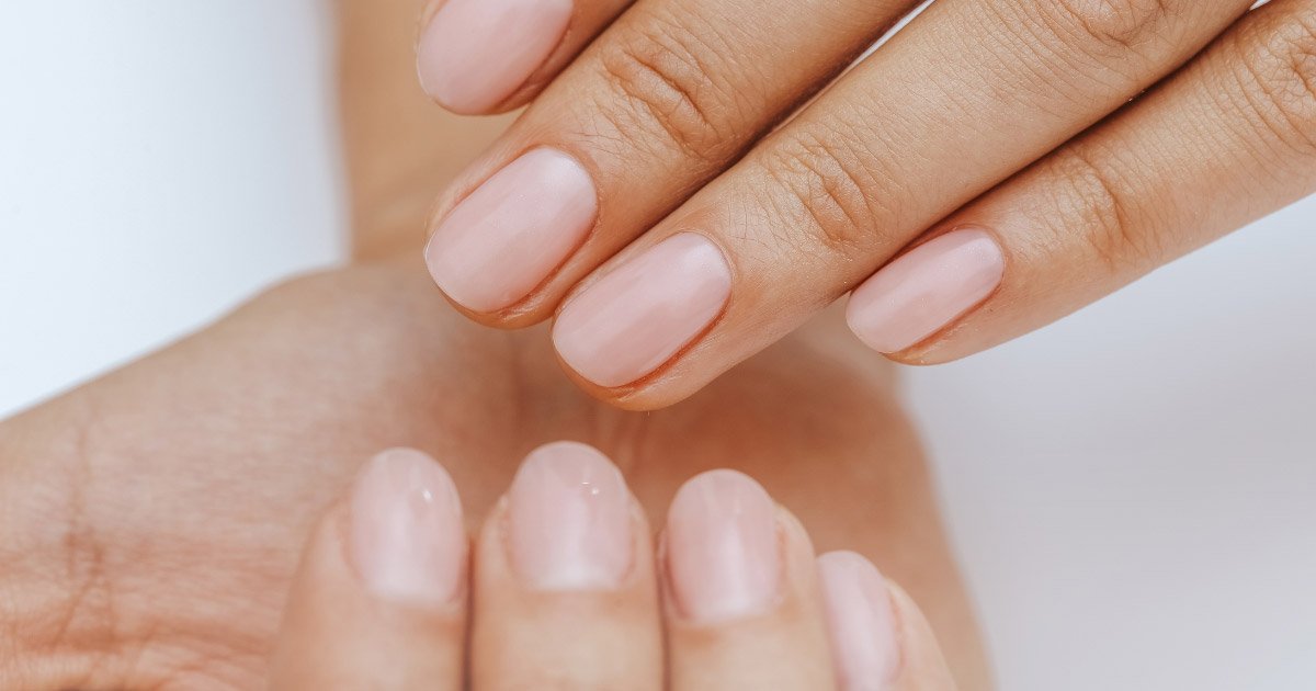 A close-up shot of two hands showing off a clean manicure with short, neatly shaped nails painted in a glossy, semi-sheer pale pink colour. 